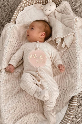 Baby in a basket lying on a knitted blanket wearing a knitted romper with knitted booties and a knitted bunny comforter, with a one month plaque