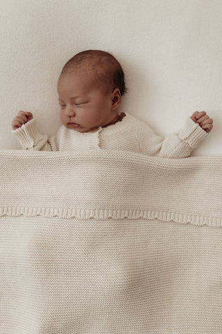 Sleeping baby wearing beige knitted romper and tucked in under a beige knitted blanket with a scalloped edge.