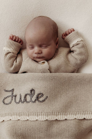 Sleeping baby wearing neutral knitted romper with matching knitted blanket with name embroidered: Jude