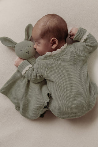 Sleeping baby wearing sage knitted romper cuddling sage green bunny comforter