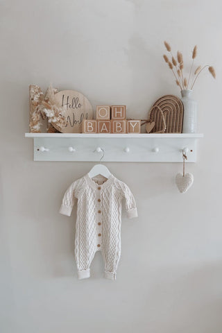 Oat cable-knit baby romper hanging in a nursery. Image also shows a shelf of blocks saying 'hello world' and 'oh baby'