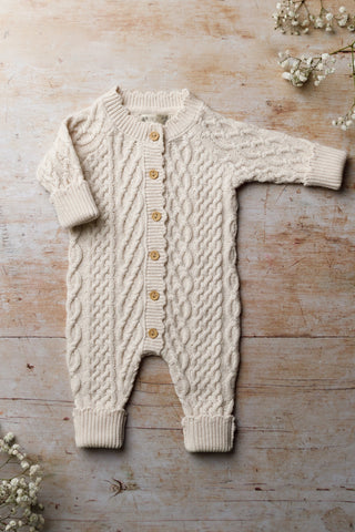 Cable-knit organic cotton baby romper photographed on a wooden background and showing the roll sleeves  and legs that can be adjusted as the baby grows