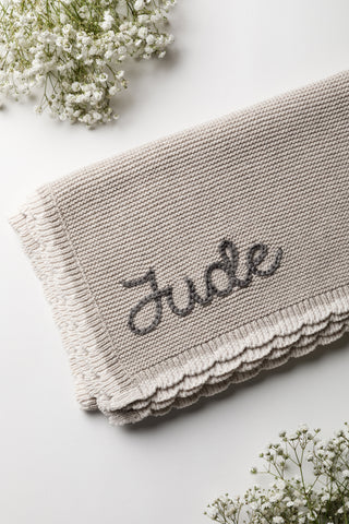 Grey knitted baby blanket with cream scalloped edge and dark grey embroidered name: Jude