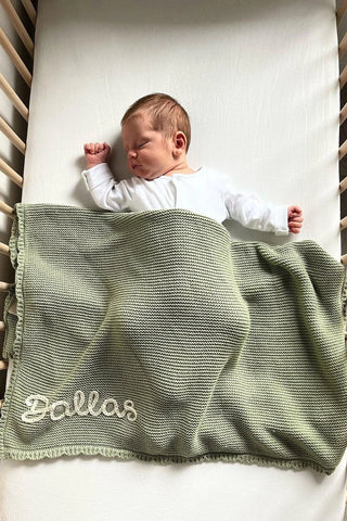 Sleeping baby in cot on white sheet with sage green knitted embroidered 'Dallas' name blanket