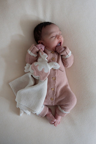 Yawning baby wearing knitted pink romper cuddling a knitted stork comforter