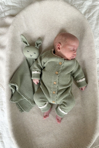 Sleeping newborn baby in basket lying on beige blanket wearing sage green knitted romper, with sage green knitted bunny comforter