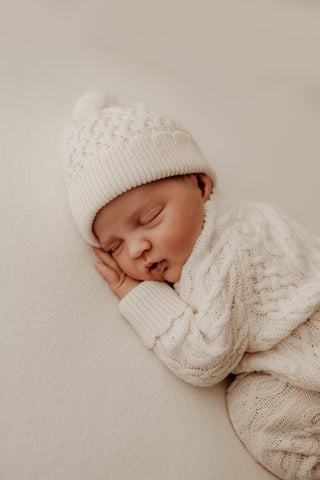 Sleeping baby lying on their front wearing cream cable knit romper and hat