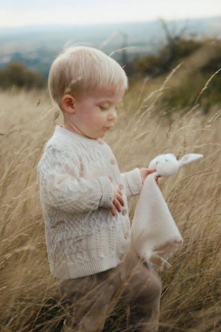Toddler Walking through field of long grass holding a knitted bunny and wearing Organic Cotton Baby Cardigan 