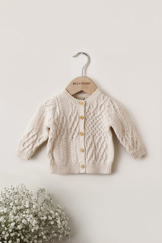 Organic Cotton Cable Knit Baby Cardigan on a hanger on a white wall with Gypsophilia in the corner