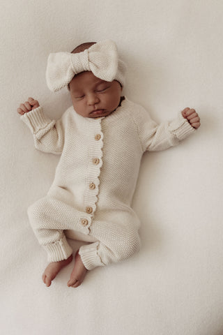 Sleeping baby wearing a neutral knitted baby headband with bow, and a button-up romper