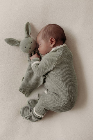 Sleeping baby wearing sage green knitted romper cuddling sage green knitted bunny comforter