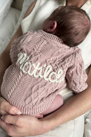 Baby in mother's arms wearing a knitted pink cable cardigan with embroidered name 'Matilda' on the back