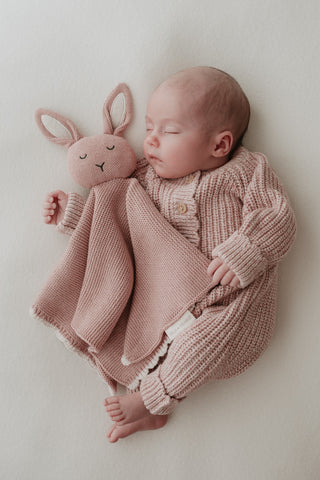 Sleeping baby wearing a light pink knitted romper cuddling a pink knitted bunny comforter with cream ears