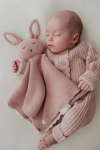 Sleeping baby wearing a knitted pink romper cuddling a pink bunny comforter
