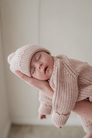 Sleeping baby being held up wearing a pink pompom hat and matching knitted outfit
