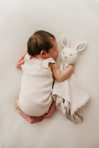 Baby lying on their front wearing a neutral knitted romper cuddling a neutral knitted bunny comforter 