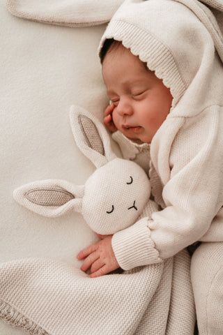 Baby wearing a neutral knitted bunny suit cuddling a neutral knitted bunny comforter 