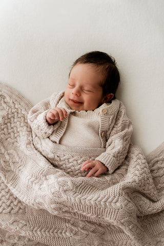 Baby smiling under a beige cable knit blanket wearing a matching cardigan