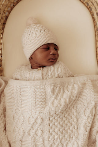 Baby in moses basket wearing knitted hat and cardigan with a cable knit blanket