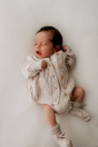 Baby wearing a knitted grey cable knit cardigan, beige romper, and grey booties
