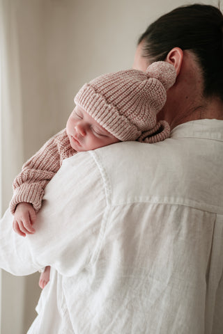 Baby sleeping on mother's shoulder wearing a light pink knitted pom pom hat