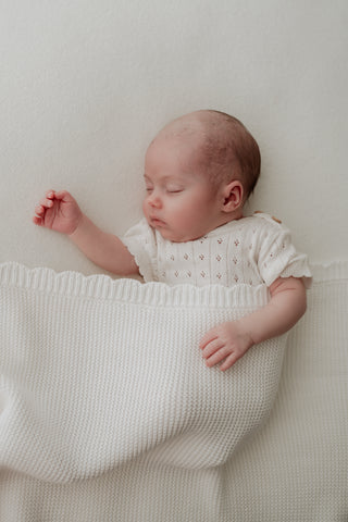 Sleeping baby wearing white pointelle bodysuit with a white scallop edge baby blanket