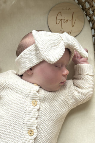 sleeping baby wearing knitted bow headband and romper, lying next to 'it's a girl' plaque 