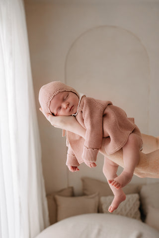 Sleeping baby being lifted up wearing a pink knitted bonnet and cardigan