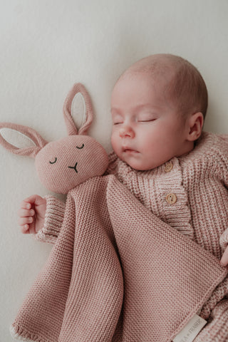 Sleeping baby wearing a pink knitted romper cuddling a pink bunny comforter
