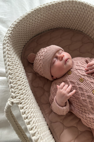 Baby in moses basket wearing pink knitted cable hat and romper