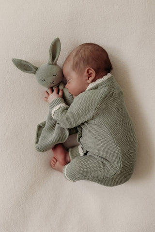 Sleeping baby lying on their side wearing a sage green knitted romper and cuddling a sage green knitted bunny comforter