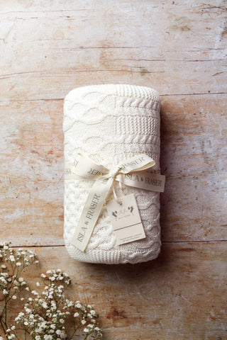 White Cable Knit Baby Blanket tied in an Isla & Fraser ribbon on a wood backdrop