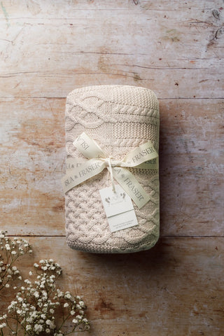 Cable Knit Baby Blanket tied with an Isla and Fraser ribbon on a wood backdrop