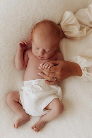 Sleeping baby on a bed with her hand and head being cradled by her mother. Baby is wearing white knitted baby bloomers 