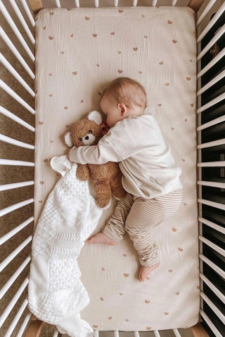 Sleeping baby in cot cuddling a teddy bear and a white knitted baby blanket