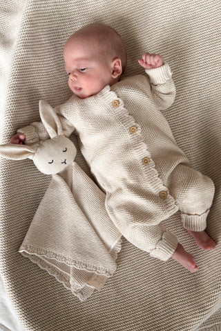Baby lying on beige knitted blanket wearing beige knitted romper with a knitted bunny comforter