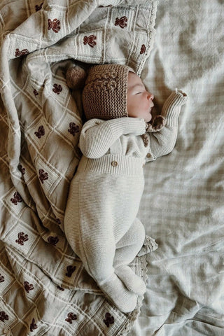 Sleeping baby on check sheets wearing knitted bonnet and wearing knitted jumper and dungarees