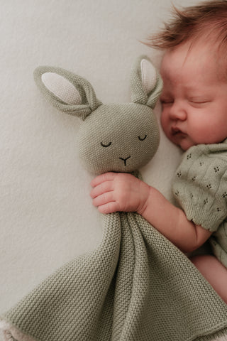 Sleeping baby wearing a sage green knitted bodysuit holding a sage green bunny comforter with cream embroidered ears 
