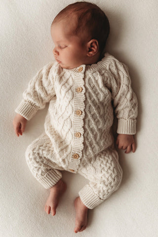 A sleeping newborn wearing the cable-knit romper in neutral beige organic cotton. Wooden buttons are shown.