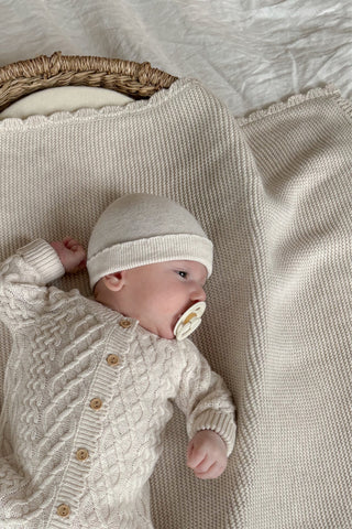 Baby with dummy in basket lying on knitted blanket with knitted hat and romper