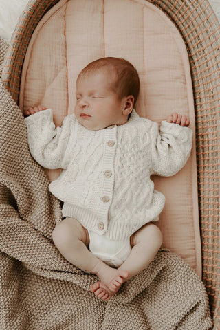 Sleeping baby in moses basket on peach basket liner with brown blanket and wearing neutral cable knit cardigan