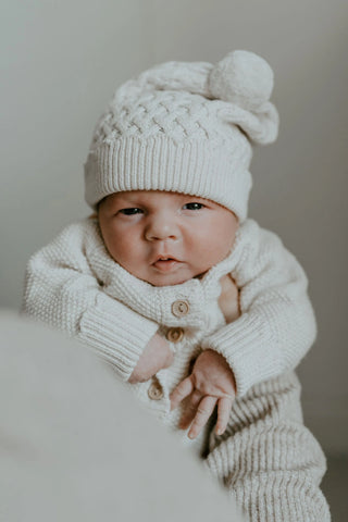 Baby being held up wearing knitted pom pom hat and knitted romper