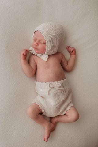 Baby wearing knitted neutral bonnet and bloomers with bow and scalloped edge