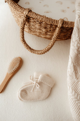 Knitted baby booties with a bow next to a baby hairbrush and baby moses basket with a cable knit blanket