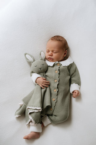 Baby wearing a knitted green outfit with white collar and cuffs and cuddling a green knitted bunny with white ears and scalloped edging