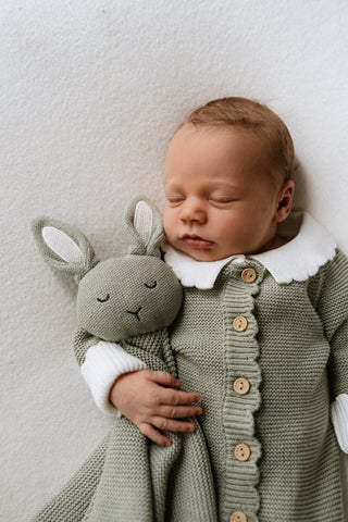 Baby wearing a knitted green outfit with white collar and cuffs and cuddling a green knitted bunny with white ears 