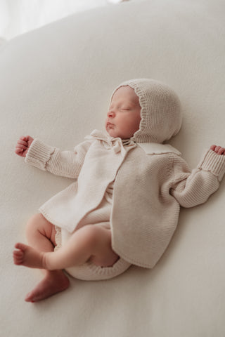 Sleeping baby wearing neutral knitted bonnet, cardigan and bloomers