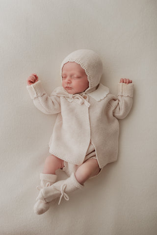Sleeping baby wearing neutral knitted bonnet, cardigan, and booties 