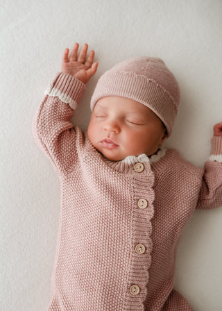 Sleeping baby wearing pink knitted hat and romper with buttons and cream scalloped edge