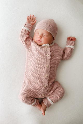 Sleeping baby wearing pink knitted hat and romper with buttons and cream scalloped edge